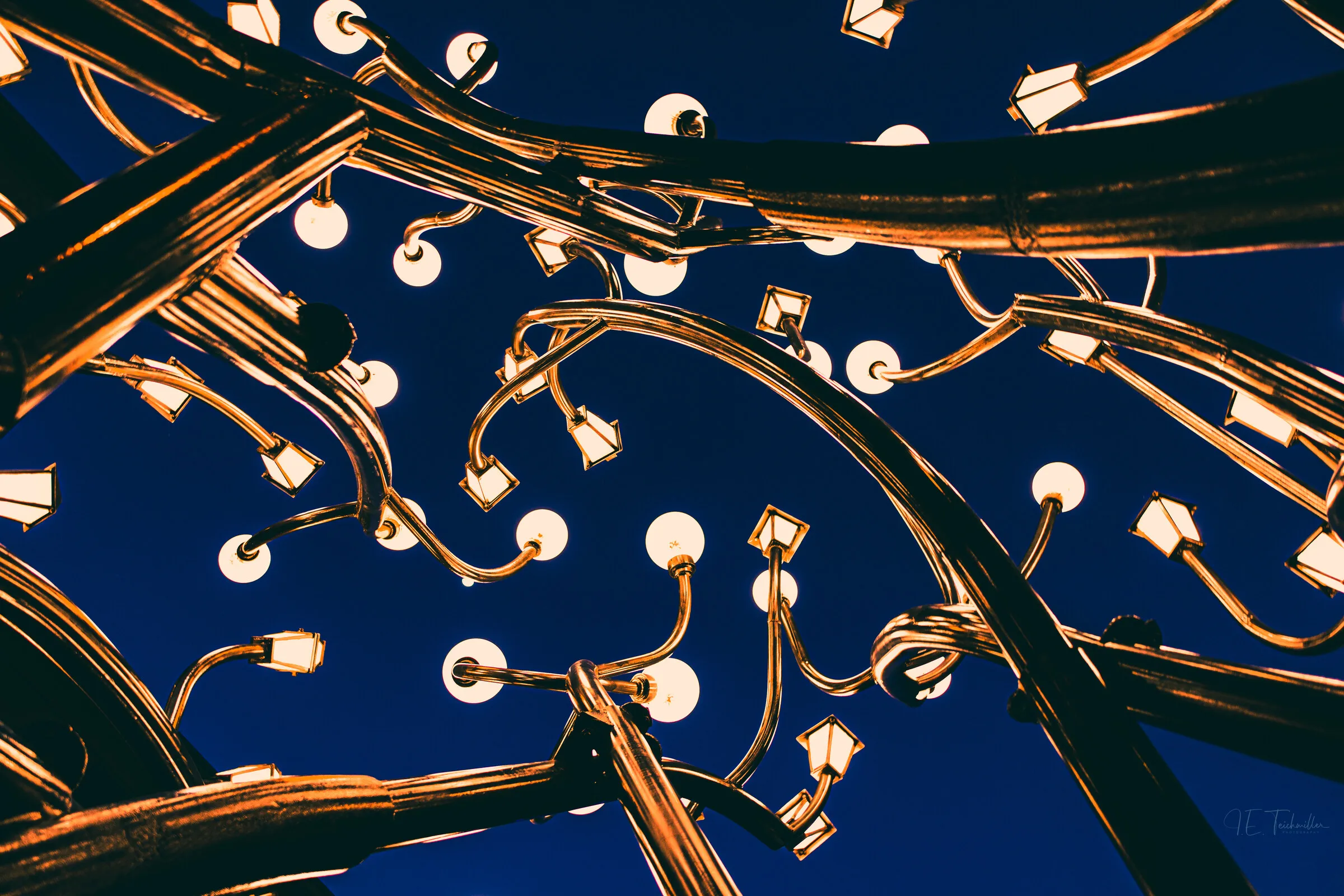 Upward view into a sculptural light installation of scrolling brass arms studded with glowing globe lamps against a deep blue dusk sky.