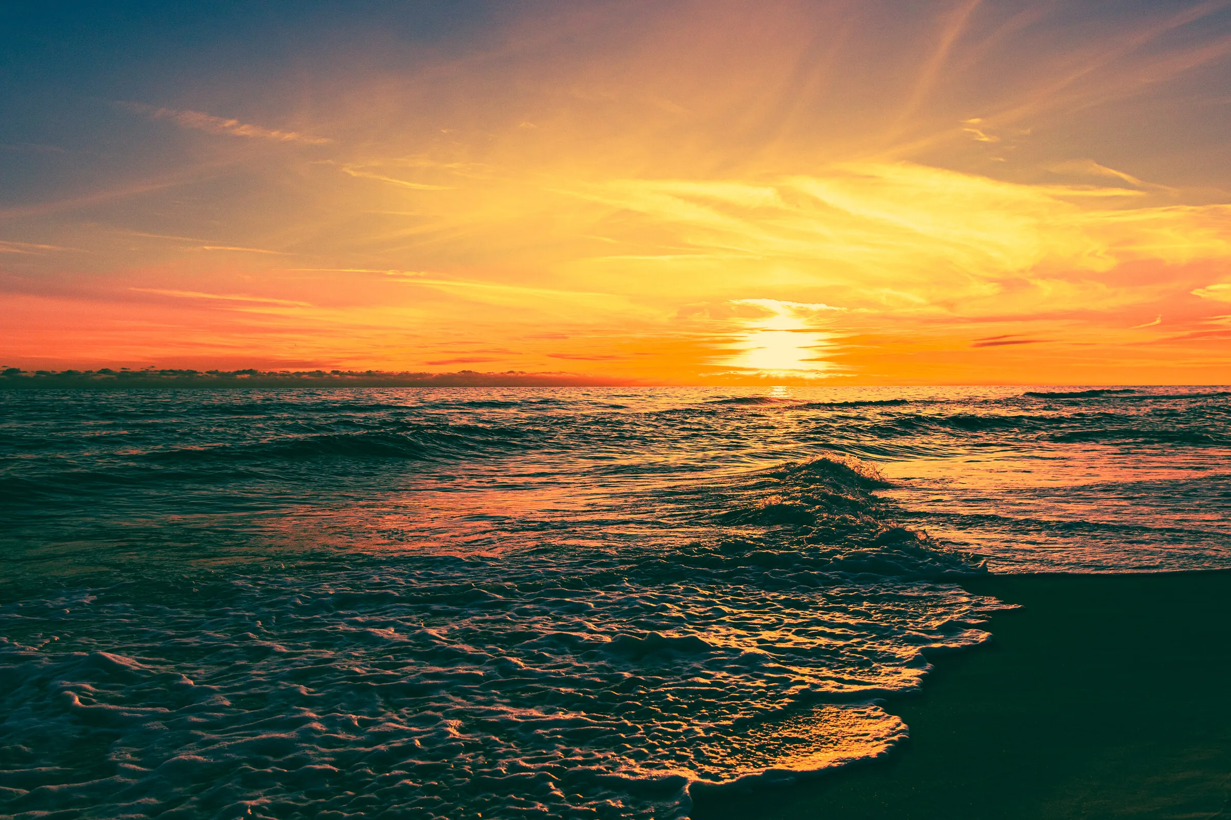 Ocean waves rolling toward shore beneath a banded gold-and-teal beach sunset.