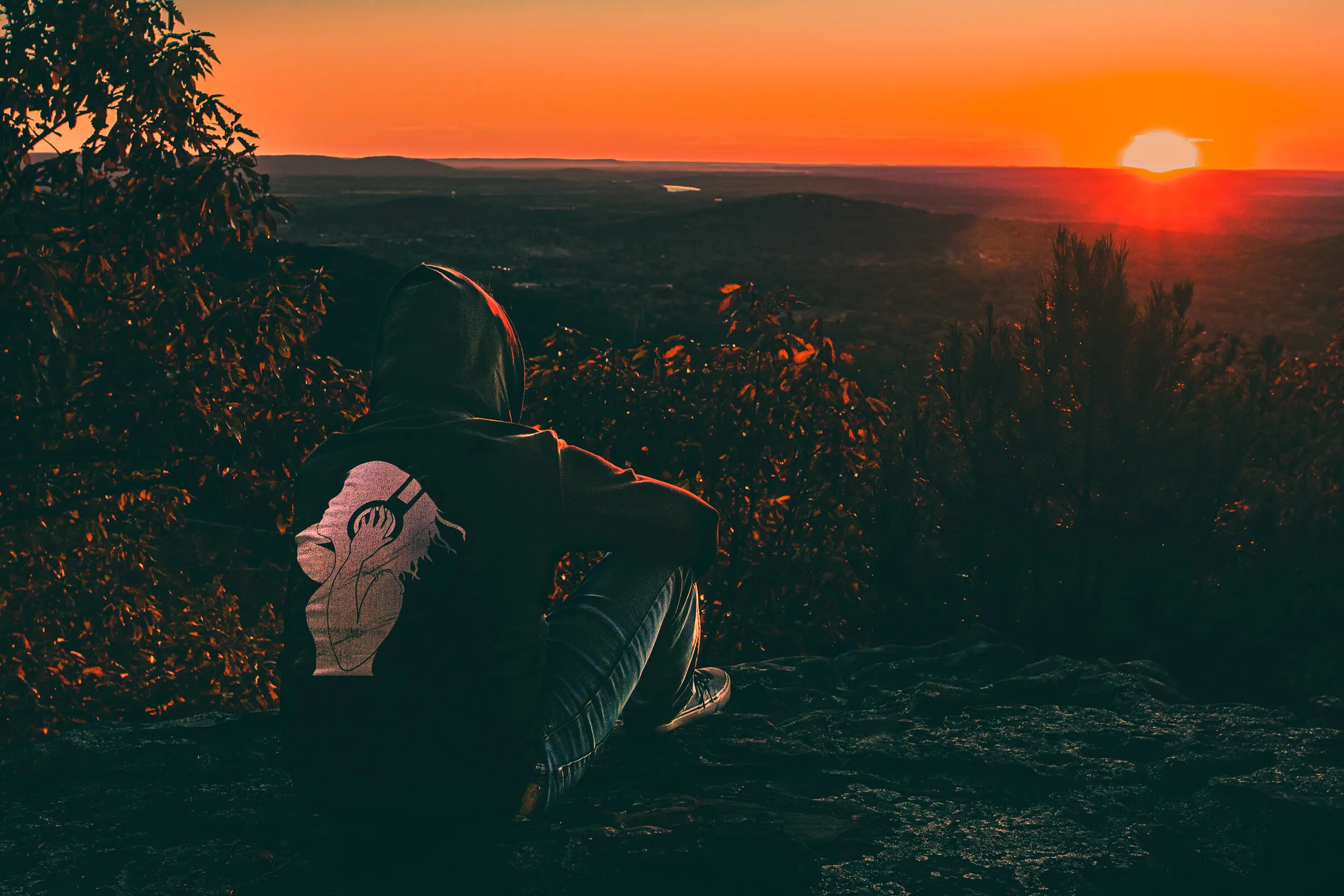 A hooded figure seen from behind, seated on a bluff, watching a fiery sun set over a hazy valley.