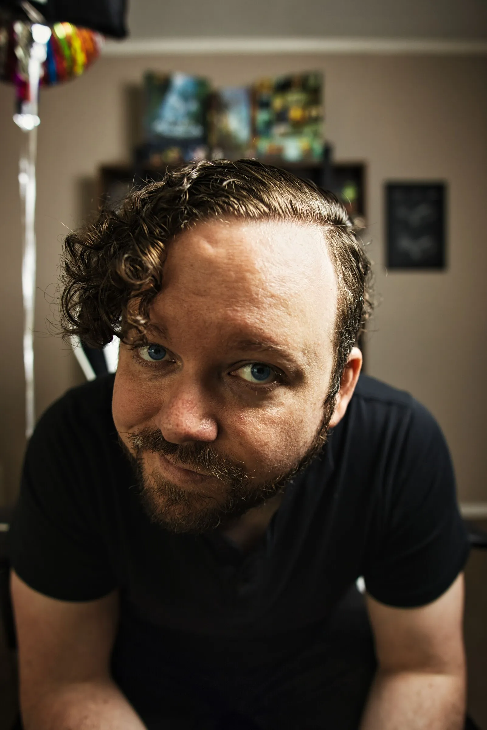 Close indoor portrait of a curly-haired, bearded man in a black shirt looking up at the camera.