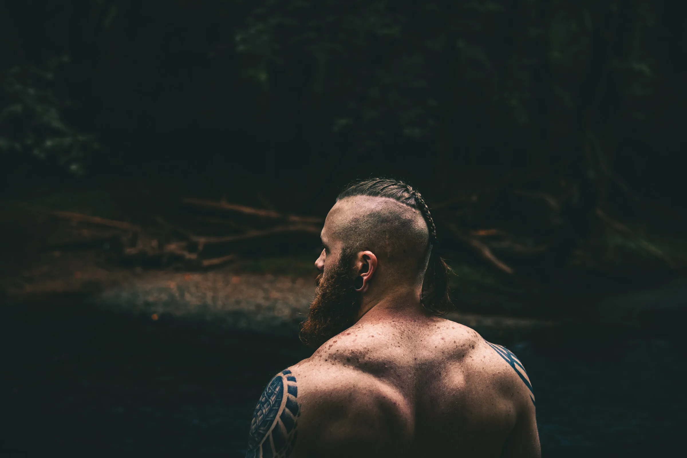 A bearded man with a braided undercut and shoulder tattoos, seen from behind facing a dark forest creek.