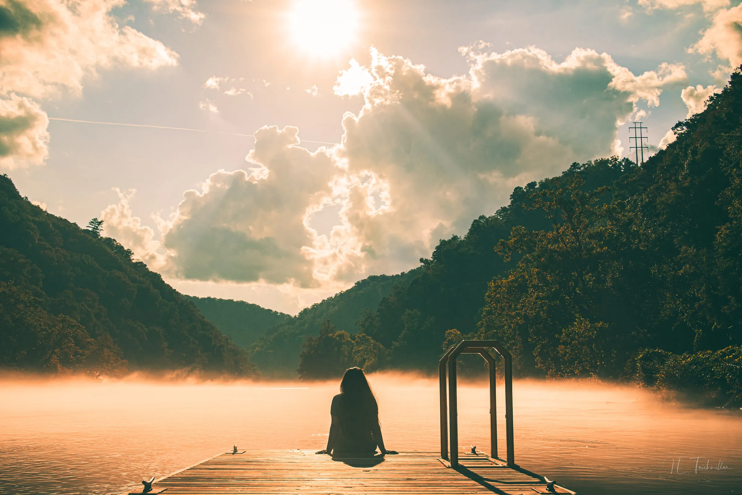 A long-haired person seated on a dock from behind, looking out over a misty lake as sunbeams break through the clouds.